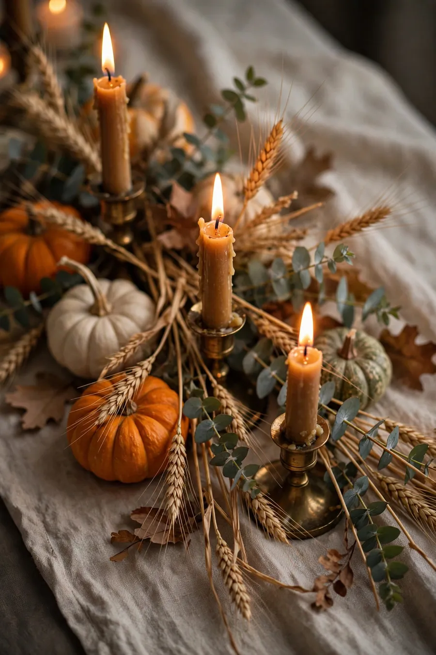 Friendsgiving table decor detail: lit amber taper candles in brass holders with small pumpkins, dried wheat and eucalyptus on a linen table runner